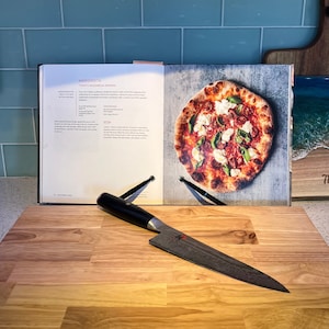 May include: A wooden chopping board with a black chef's knife on top. A cookbook with a recipe for "Margherita" pizza is open on the board. The pizza is pictured on the page, with a red sauce base, mozzarella cheese, and basil.