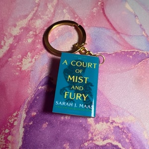 Puede incluir: Un llavero dorado con un dije de libro azul con el título "A Court of Mist and Fury" de Sarah J. Maas.