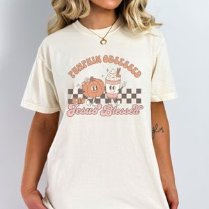 May include: Cream-colored t-shirt with the words "PUMPKIN OBSESSED" and "Jesus Blessed" in a retro font. The graphic features a cartoon pumpkin and a coffee cup with whipped cream, set against a checkered background.