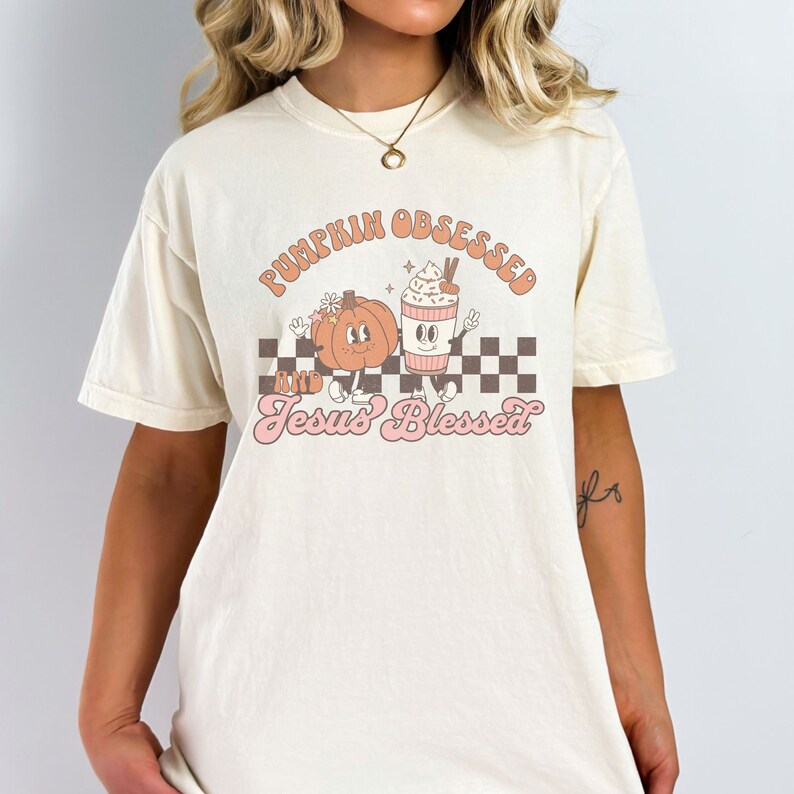 May include: Cream-colored t-shirt with the words "PUMPKIN OBSESSED" and "Jesus Blessed" in a retro font. The graphic features a cartoon pumpkin and a coffee cup with whipped cream, set against a checkered background.