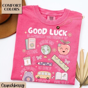 May include: A pink t-shirt with the words "GOOD LUCK" and other motivational phrases and illustrations. The shirt features a crew neck and the Comfort Colors brand label. A gold necklace is draped across the shirt.