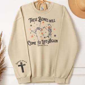 May include: A beige sweatshirt with a graphic print of two skeletons dancing with stars and the text "These bones will come to life again Ezekiel 37:4-5".
