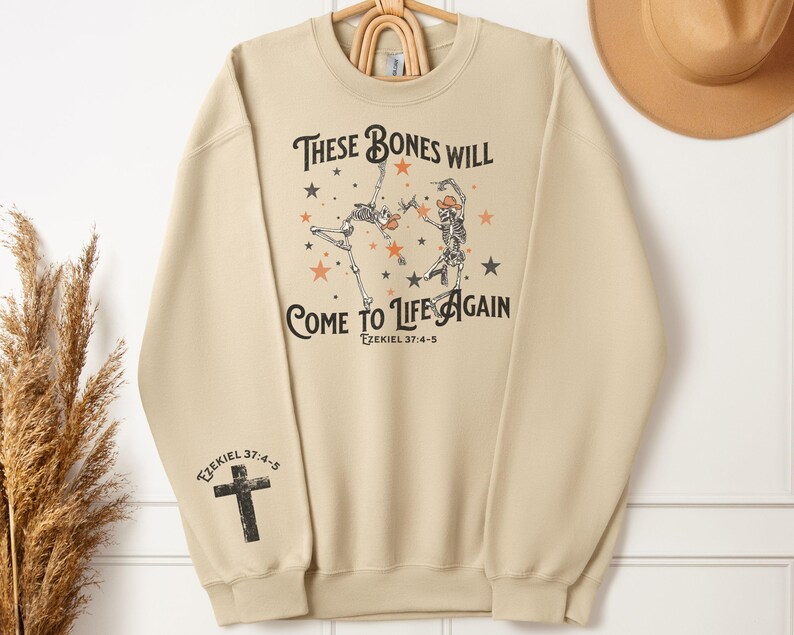 May include: A beige sweatshirt with a graphic print of two skeletons dancing with stars and the text "These bones will come to life again Ezekiel 37:4-5".