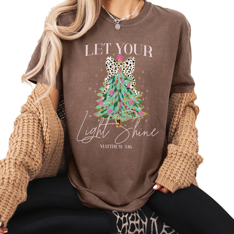 May include: Brown t-shirt with the text "LET YOUR LIGHT SHINE" and a Christmas tree design. The tree is teal with pink ornaments and a black and white bow. The text "Matthew 5:16" is below the tree.