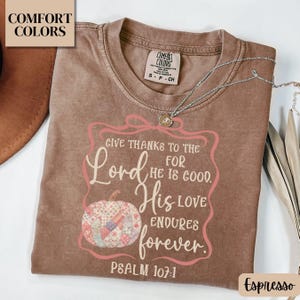 May include: Espresso-colored t-shirt with the text "Give thanks to the Lord for He is good. His love endures forever. Psalm 107:1." A decorative pumpkin is featured in the design. A silver necklace with a pendant is on the shirt.