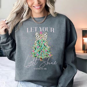 May include: A gray sweatshirt with a Christmas tree design. The tree is teal with pink and gold accents, and a black and white bow. The text "LET YOUR LIGHT SHINE" and "MATTHEW 5:16" are printed on the sweatshirt.