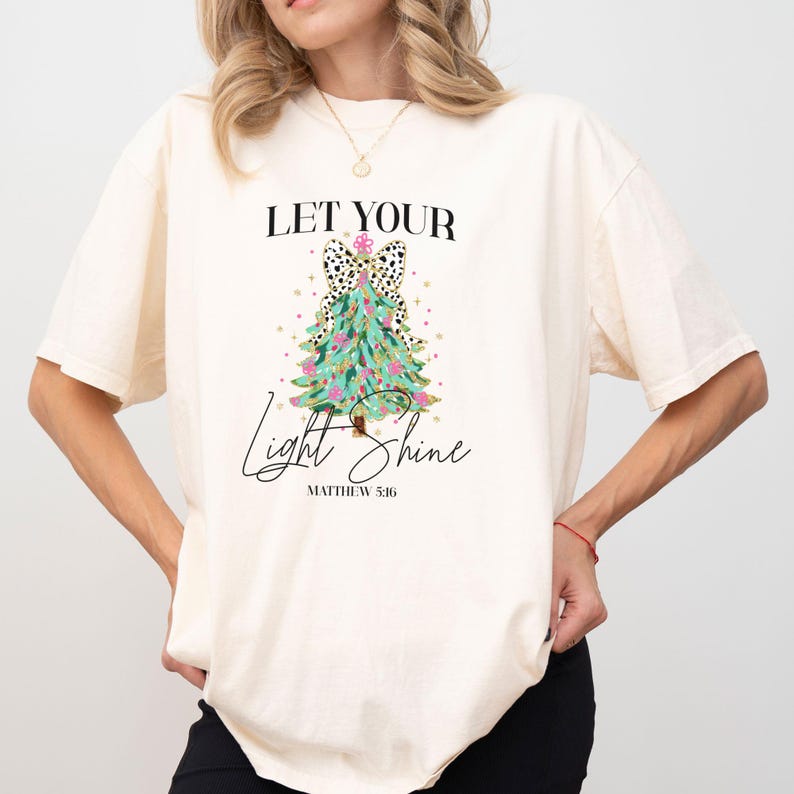 May include: Cream-colored t-shirt featuring the text "LET YOUR Light Shine" above a Christmas tree design. The tree is decorated with teal and pink accents, and a black and white spotted bow. The text "Matthew 5:16" is below the design.