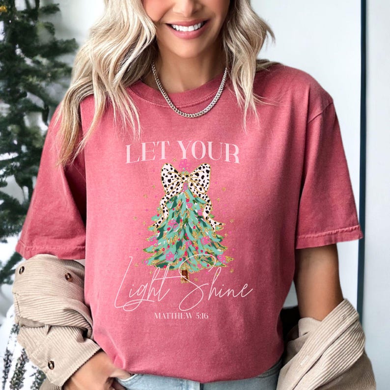 May include: A red-pink t-shirt with a Christmas tree graphic. The tree is decorated with a leopard print bow, gold accents, and the words "Let Your Light Shine" and "Matthew 5:16".