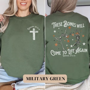 May include: A military green sweatshirt with a cross on the front and a graphic of two skeletons dancing with the text "These bones will come to life again" and "Ezekiel 37:4-5" on the back.