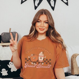 May include: A burnt orange t-shirt with the text "Pumpkinn Obsessed and Jesus Blessed" in a retro font. The graphic features a cartoon pumpkin and a coffee cup. The shirt is worn by a person.