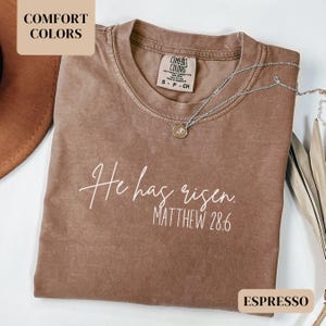 May include: A brown t-shirt with the text "He has risen. Matthew 28:6" in white lettering. The shirt is made of a soft, comfortable material and has a distressed look.