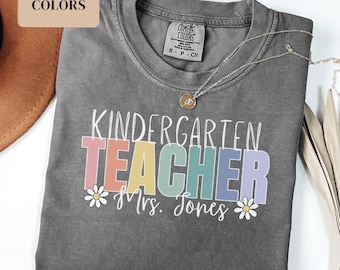 May include: A gray t-shirt with the text "KINDERGARTEN TEACHER" in rainbow colors and the text "Mrs. Jones" below. The shirt is made by Comfort Colors.