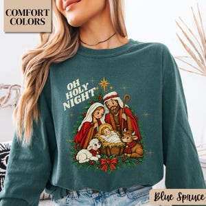 May include: A dark green crewneck sweatshirt featuring a graphic of the nativity scene with Mary, Joseph, and baby Jesus in a manger. The text reads OH HOLY NIGHT above the figures, surrounded by a wreath of pine branches, berries, and a star.
