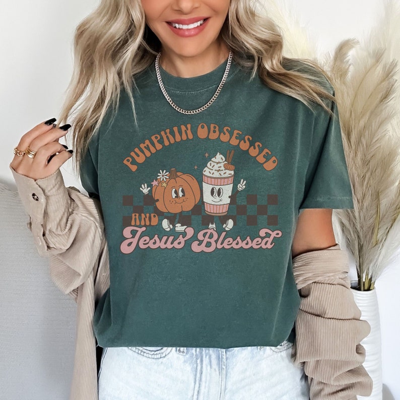 May include: Dark green t-shirt with the text "PUMPKIN OBSESSED and Jesus Blessed" in orange and pink. The graphic features a cartoon pumpkin and a coffee cup with whipped cream. The shirt has a relaxed fit.