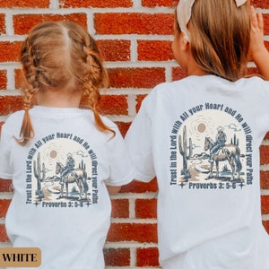 May include: Two white t-shirts with a graphic design of a cowboy riding a horse in a desert landscape. The text "Trust in the Lord with All your Heart and He will direct your Paths" is printed on the shirts. The text "Proverbs 3:5-6" is also printed on the shirts.