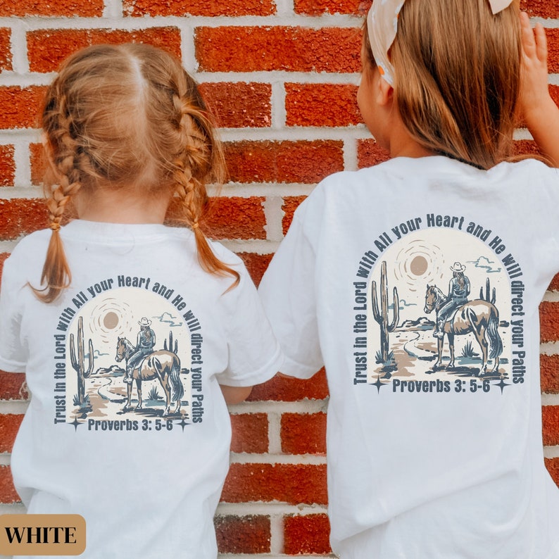 May include: Two white t-shirts with a graphic design of a cowboy riding a horse in a desert landscape. The text "Trust in the Lord with All your Heart and He will direct your Paths" is printed on the shirts. The text "Proverbs 3:5-6" is also printed on the shirts.