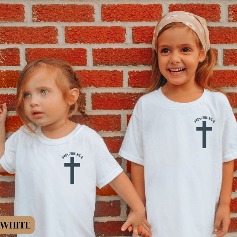 May include: Two children wearing white t-shirts with a black cross and the text "Proverbs 3:5-6" printed on them. The children are standing in front of a red brick wall. The child on the left is looking at the camera, while the child on the right is smiling and looking to the side.