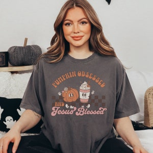 May include: Dark gray t-shirt with the text "PUMPKIN OBSESSED and Jesus Blessed" in orange and pink. The shirt features cartoon pumpkins and a coffee cup with whipped cream. The shirt is a relaxed fit.