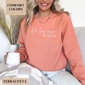 May include: A terracotta colored sweatshirt with the text "He has risen. Matthew 28:6" in white.