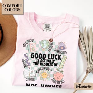 May include: A light pink t-shirt with the words "Good Luck is Actually the Results of Making Good Choices" and other school-themed graphics. The shirt also has the words "Comfort Colors" printed on it.