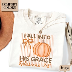 May include: Ivory-colored t-shirt with a graphic featuring a cross, pumpkin, and the text "FALL INTO HIS GRACE Ephesians 2:8." The design is in shades of orange and brown. The shirt has a Comfort Colors label.