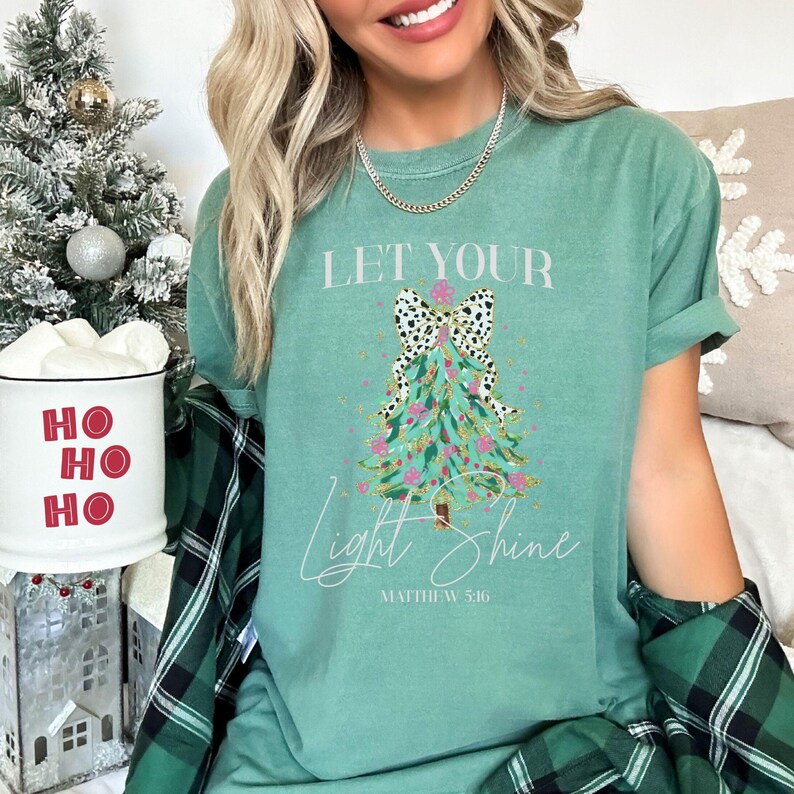 May include: A seafoam green t-shirt with the text "LET YOUR Light Shine" and a Christmas tree design. The tree is decorated with pink and gold accents and a black and white bow. The shirt also includes the Bible verse Matthew 5:16.
