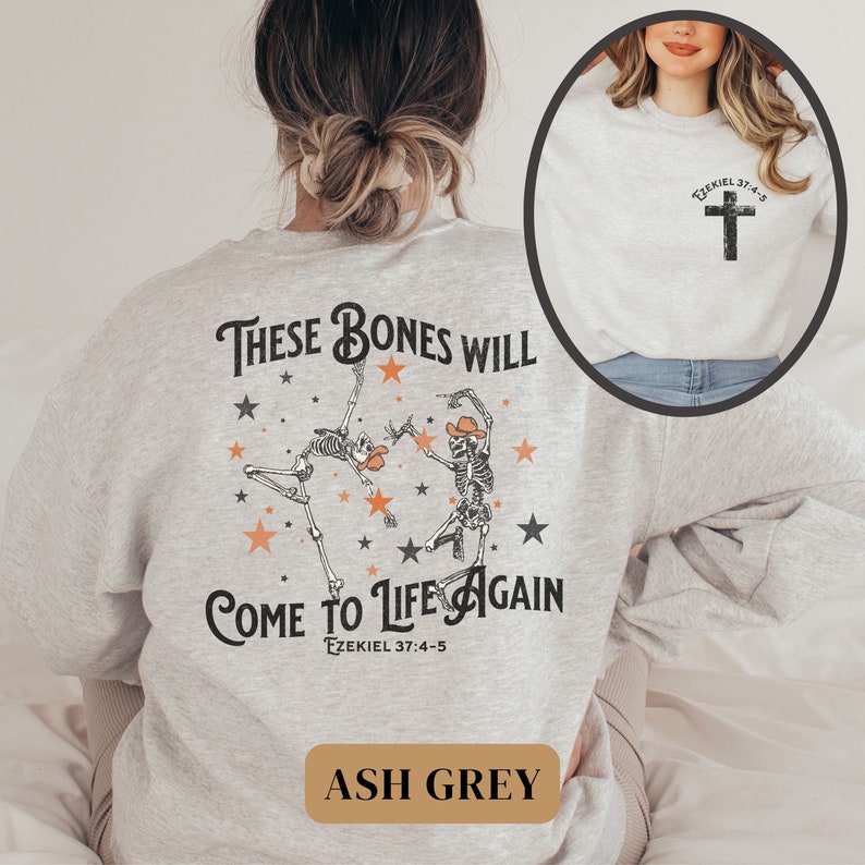 May include: A light grey sweatshirt with a graphic of two skeletons dancing with stars and the text "These bones will come to life again Ezekiel 37:4-5". The sweatshirt also has the text "Ezekiel 37:4-5" and a cross on the front. The sweatshirt is labeled "Ash Grey".