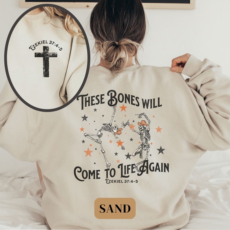 May include: A beige sweatshirt with a graphic print of two skeletons dancing with stars and the text "These bones will come to life again Ezekiel 37:4-5". The back of the sweatshirt has a cross with the text "Ezekiel 37:4-5".