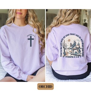 May include: A lavender sweatshirt with a cross and the text "Proverbs 3:5-6" on the front. The back of the sweatshirt features a cowboy riding a horse in a desert landscape with the text "Trust in the Lord with All your Heart and He will direct your Paths" and "Proverbs 3:5-6".