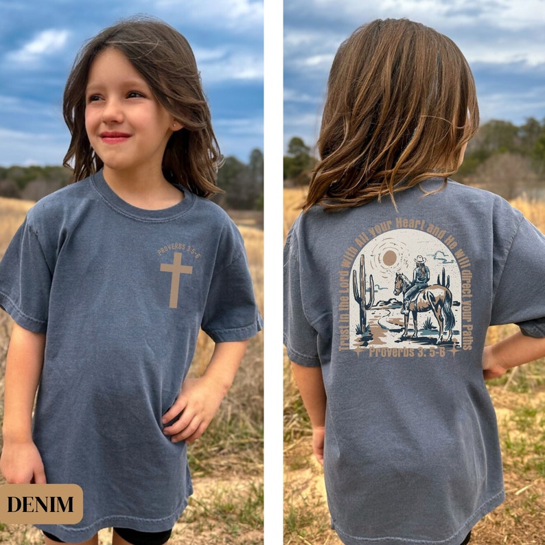 May include: A denim blue t-shirt with a brown cross and the text "Proverbs 3:5-6" on the front. The back of the shirt features a brown illustration of a cowboy riding a horse in a desert landscape with the text "Trust in the Lord with all your heart and He will direct your paths" and "Proverbs 3:5-6".