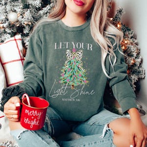 May include: Dark green sweatshirt with the text "Let Your Light Shine" and a Christmas tree design. The tree has a dalmatian print bow and is decorated with pink and gold accents. A red mug with "merry + bright" is held in the person's hand.