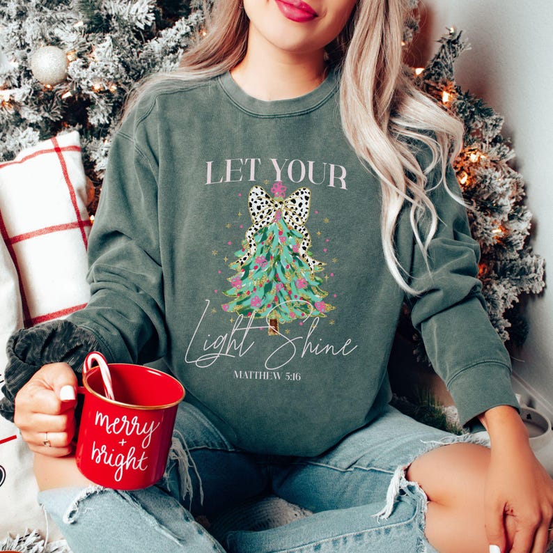 May include: Dark green sweatshirt with the text "Let Your Light Shine" and a Christmas tree design. The tree has a dalmatian print bow and is decorated with pink and gold accents. A red mug with "merry + bright" is held in the person's hand.