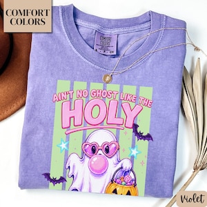 May include: A violet Comfort Colors t-shirt featuring a graphic of a ghost wearing heart-shaped pink sunglasses and blowing a bubblegum bubble. The text reads "AIN'T NO GHOST LIKE THE HOLY" above a pumpkin bucket filled with candy and bats.
