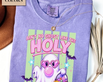 May include: A violet Comfort Colors t-shirt featuring a graphic of a ghost wearing heart-shaped pink sunglasses and blowing a bubblegum bubble. The text reads "AIN'T NO GHOST LIKE THE HOLY" above a pumpkin bucket filled with candy and bats.