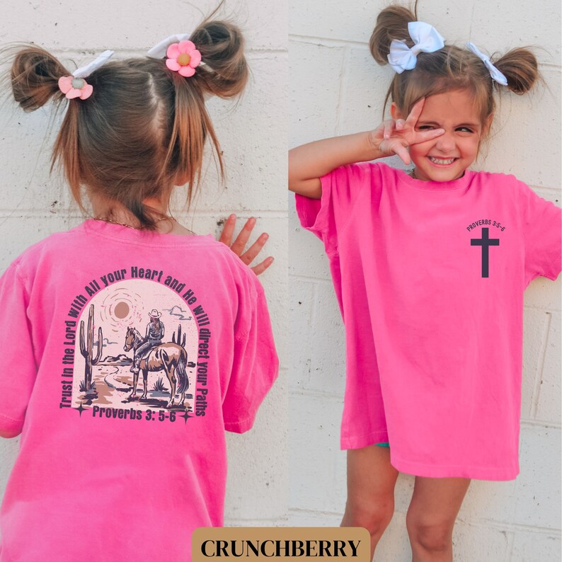 May include: Pink t-shirt with a graphic of a cowboy riding a horse in a desert landscape. The text "Trust in the Lord with All your Heart and He will direct your Paths" and "Proverbs 3:5-6" is printed on the back of the shirt. The front of the shirt has a black cross with the text "Proverbs 3:5-6".