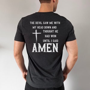 May include: A dark gray t-shirt with a white cross and the text "THE DEVIL SAW ME WITH MY HEAD DOWN AND THOUGHT HE HAD WON UNTIL I SAID AMEN".