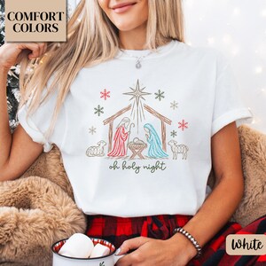 May include: A white t-shirt featuring a line art graphic of a nativity scene with a star, sheep, and the text "oh holy night" in green. The design includes red and green snowflakes surrounding the central manger scene.