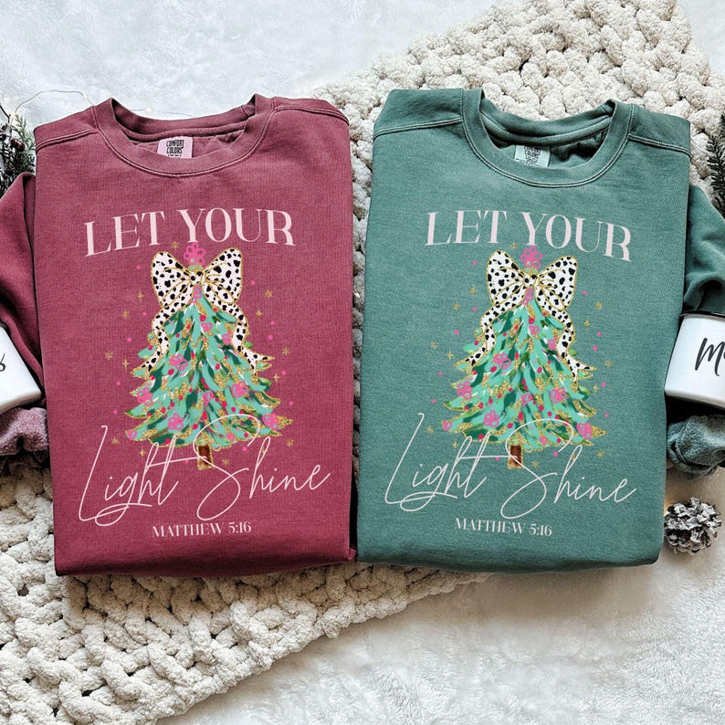 May include: Two crewneck sweatshirts, one burgundy and one teal, with a Christmas tree design. The tree has a dalmatian print bow and the text "Let Your Light Shine" with "Matthew 5:16" below.