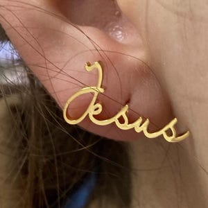 Może przedstawiać: Złoty kolczyk z napisem "Jezus" napisanym kursywą.