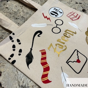 May include: Beige canvas tote bag featuring various black, red, and gold graphics. Includes a lightning bolt, glasses, a broom, a scarf, and a letter. The name "Kareen" is written in gold script. The bag has two beige handles.
