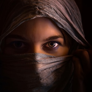 May include: A woman wearing a gray headscarf that covers most of her face, with only her eyes visible. The eyes are looking directly at the camera, and one eye has a hint of blue eyeshadow.