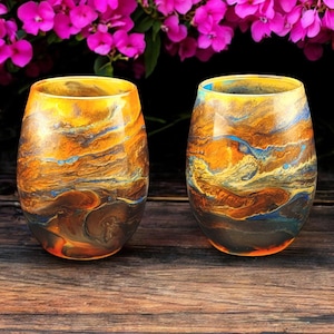 May include: Two clear glass tumblers with a swirling pattern of orange, yellow, blue, and brown paint. The glasses are sitting on a wooden surface.