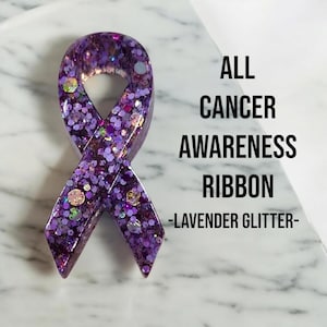 Puede incluir: Cinta de resina con purpurina morada, con forma de cinta de concienciación. El texto "ALL CANCER AWARENESS RIBBON -LAVENDER GLITTER- " está impreso debajo de la cinta.