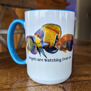 May include: White ceramic mug with a blue rim and handle. The mug features a colorful illustration of three fish with the text "Angels are watching over me".