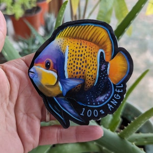 以下が含まれることがあります： 青、黄色、オレンジ、黒のカラフルな魚のステッカー。ステッカーには「100％ ANGEL」の文字が書かれています。
