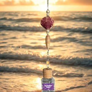 Pode incluir: Enfeite decorativo suspenso com uma concha, uma conta de madeira e uma pequena garrafa de vidro. A garrafa tem uma tampa de madeira e um rótulo que diz "lavendar haze". O enfeite é suspenso por uma corrente de prata. O fundo é uma cena de praia ao pôr do sol.