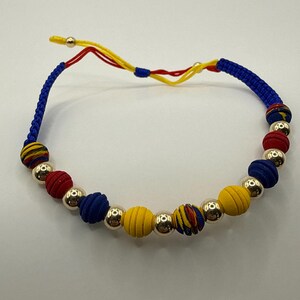 May include: A vibrant bracelet with fabric-covered beads in red, blue, and yellow, complemented by silver-coloured metal beads. The bracelet is strung on a blue cord with yellow and red detailing.
