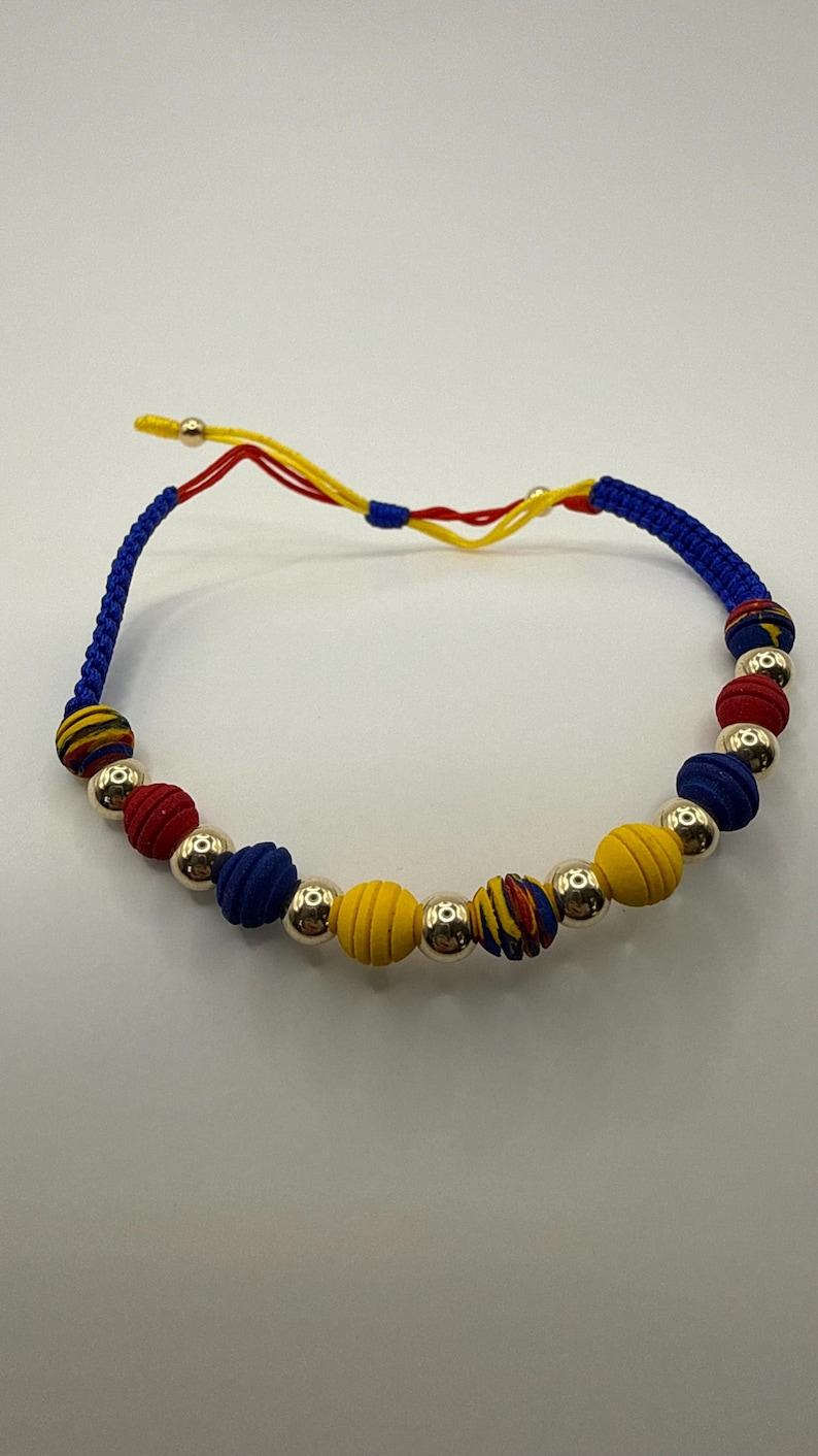 May include: A vibrant bracelet with fabric-covered beads in red, blue, and yellow, complemented by silver-coloured metal beads. The bracelet is strung on a blue cord with yellow and red detailing.