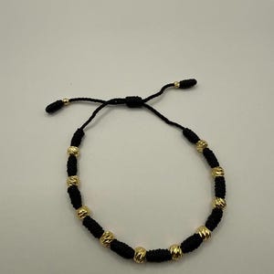 Op de afbeelding: Zwart koordarmband met goudkleurige kralen. De armband heeft een schuifknoopsluiting voor verstelbare maatvoering. De gouden kralen hebben een getextureerd, facetontwerp. De armband wordt getoond tegen een witte achtergrond.