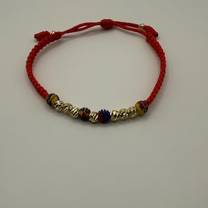 May include: Red string bracelet with gold-coloured beads and colourful accent beads. The bracelet has a sliding knot closure for adjustable sizing. The beads have a textured, ridged design.